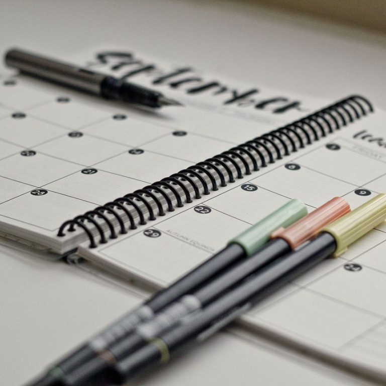 A spiral-bound planner with calendar pages and colourful pens arranged beside it.