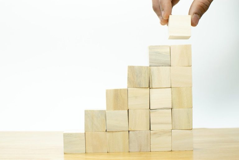 A hand places a wooden block on a step-like arrangement of stacked blocks.