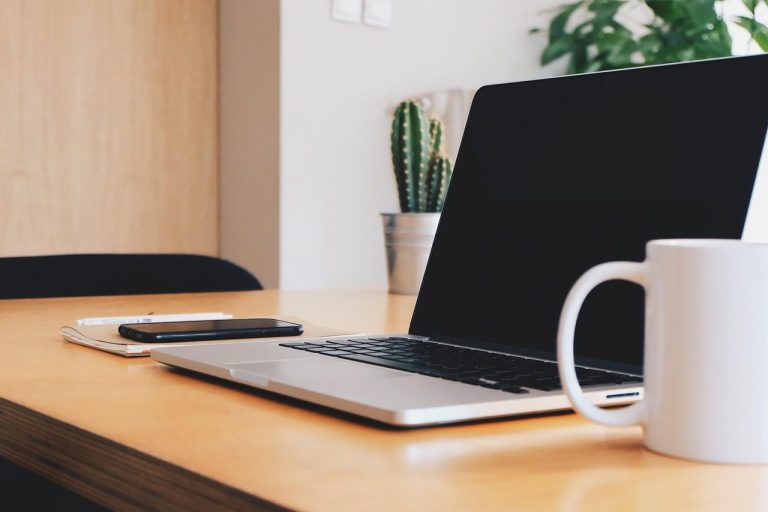 A laptop, smartphone, and mug on a clean wooden desk with plants in the background.