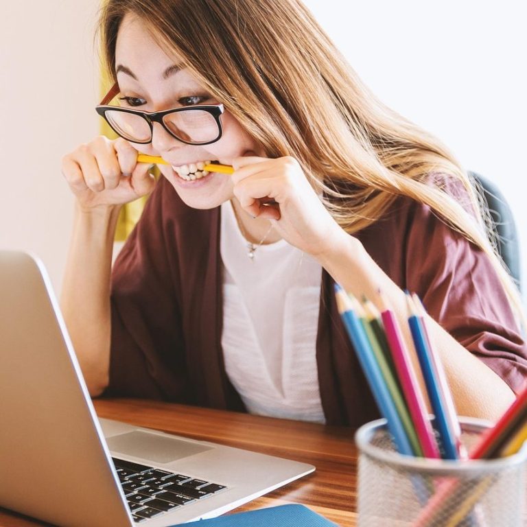 Young woman with long hair, wearing glasses, focused on a laptop while holding a pencil.
