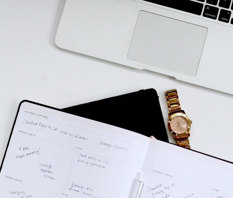 Laptop, notebook with planner notes, tablet, and a wristwatch on a white surface.